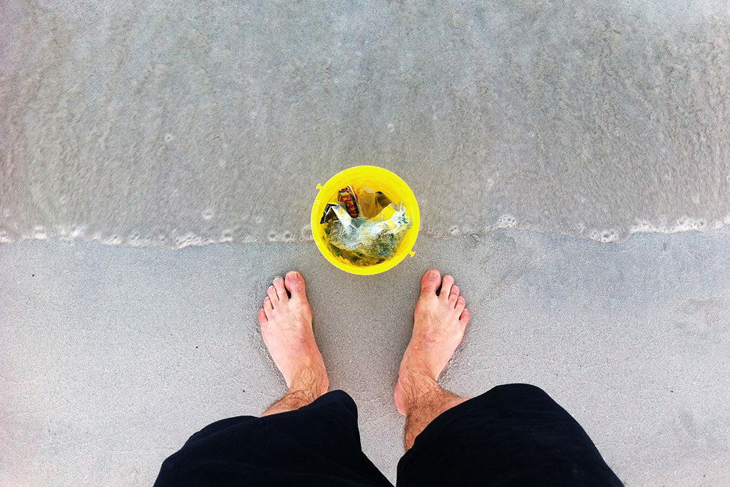Bare feet next to a yellow bucket filled with beach plastic on the sand
