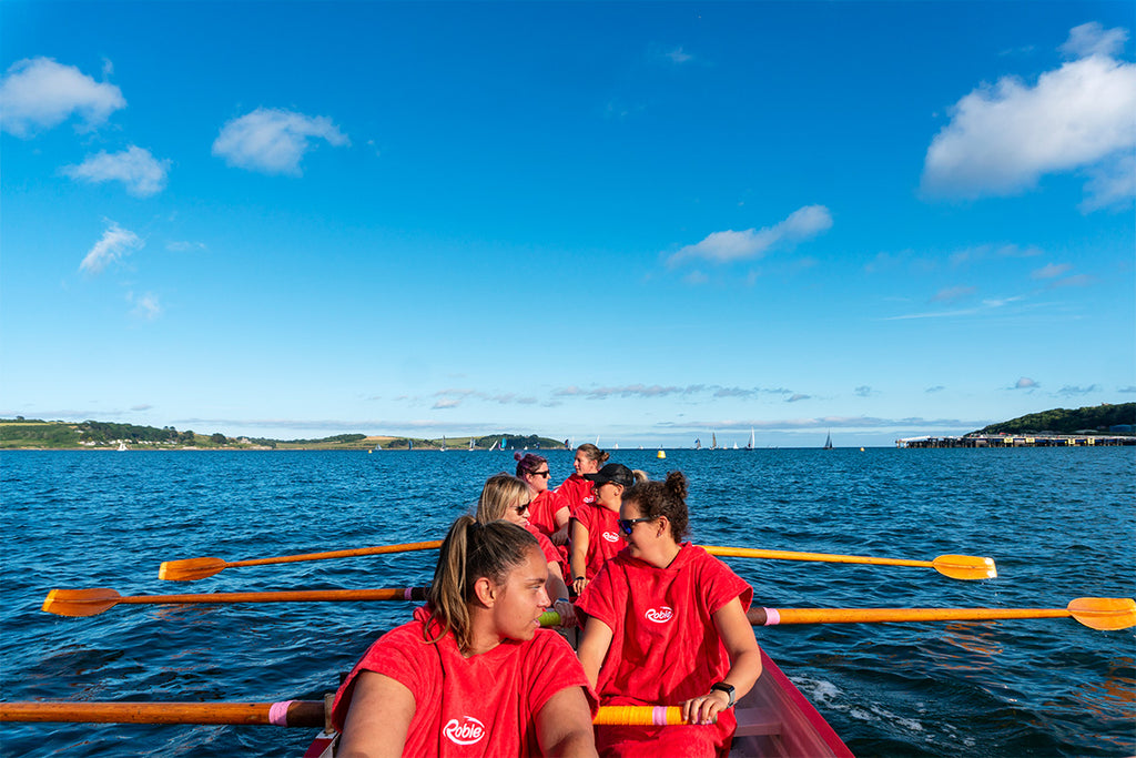 women wearing red robies rowing a gig boat out to sea in the sunshine