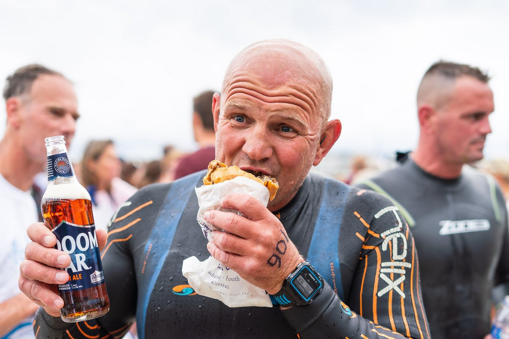 bald man in wetsuit eats a pasty with pint in hand