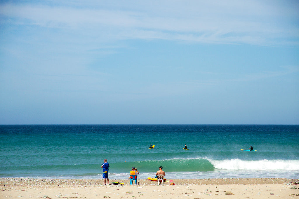 people sit on a sunny beach watching surfers in a bright blue sea